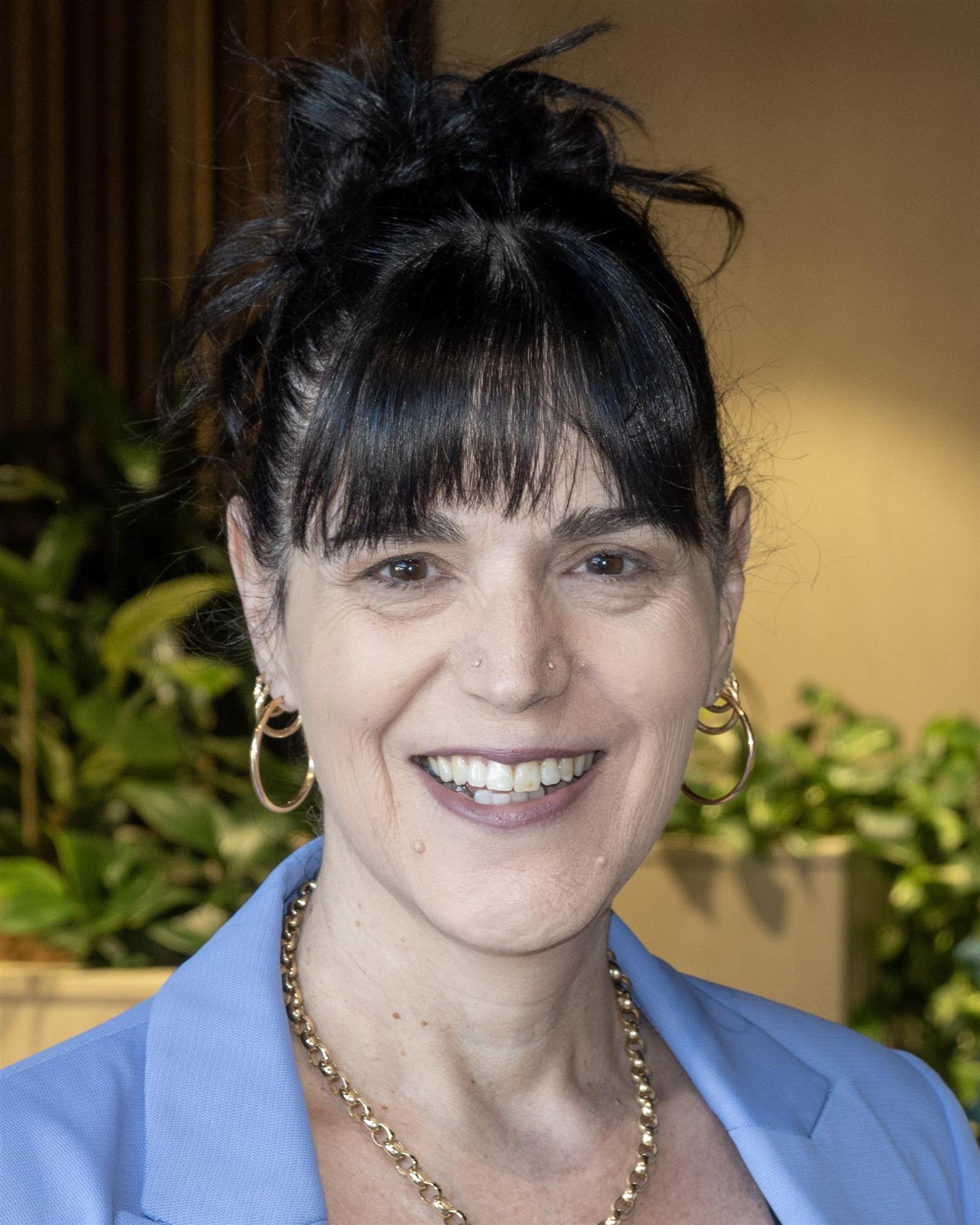 A head‑and‑shoulders portrait of a person facing the camera, with dark hair tied up and a fringe, wearing hoop earrings, a gold chain necklace and a light blue jacket. Green plants are visible in the softly lit background.