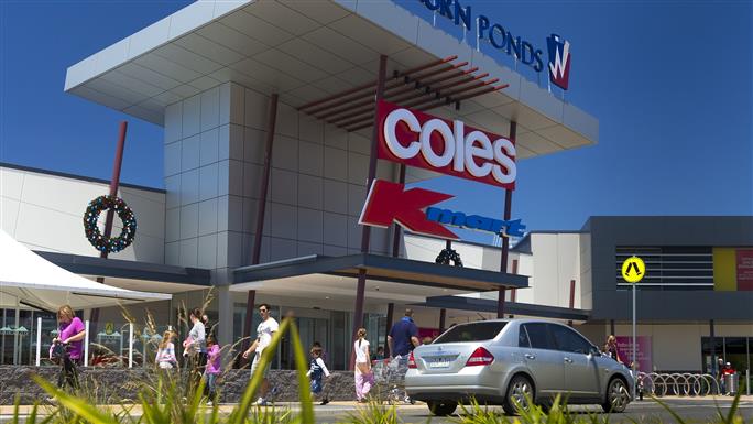 Exterior of a Coles supermarket at Waurn Ponds shopping centre, showing the store entrance, Coles signage, a covered walkway, and people walking past with parked cars nearby.