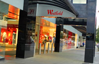 Exterior of a Westfield shopping centre entrance, showing glass storefronts, illuminated retail displays, and a covered pedestrian walkway.