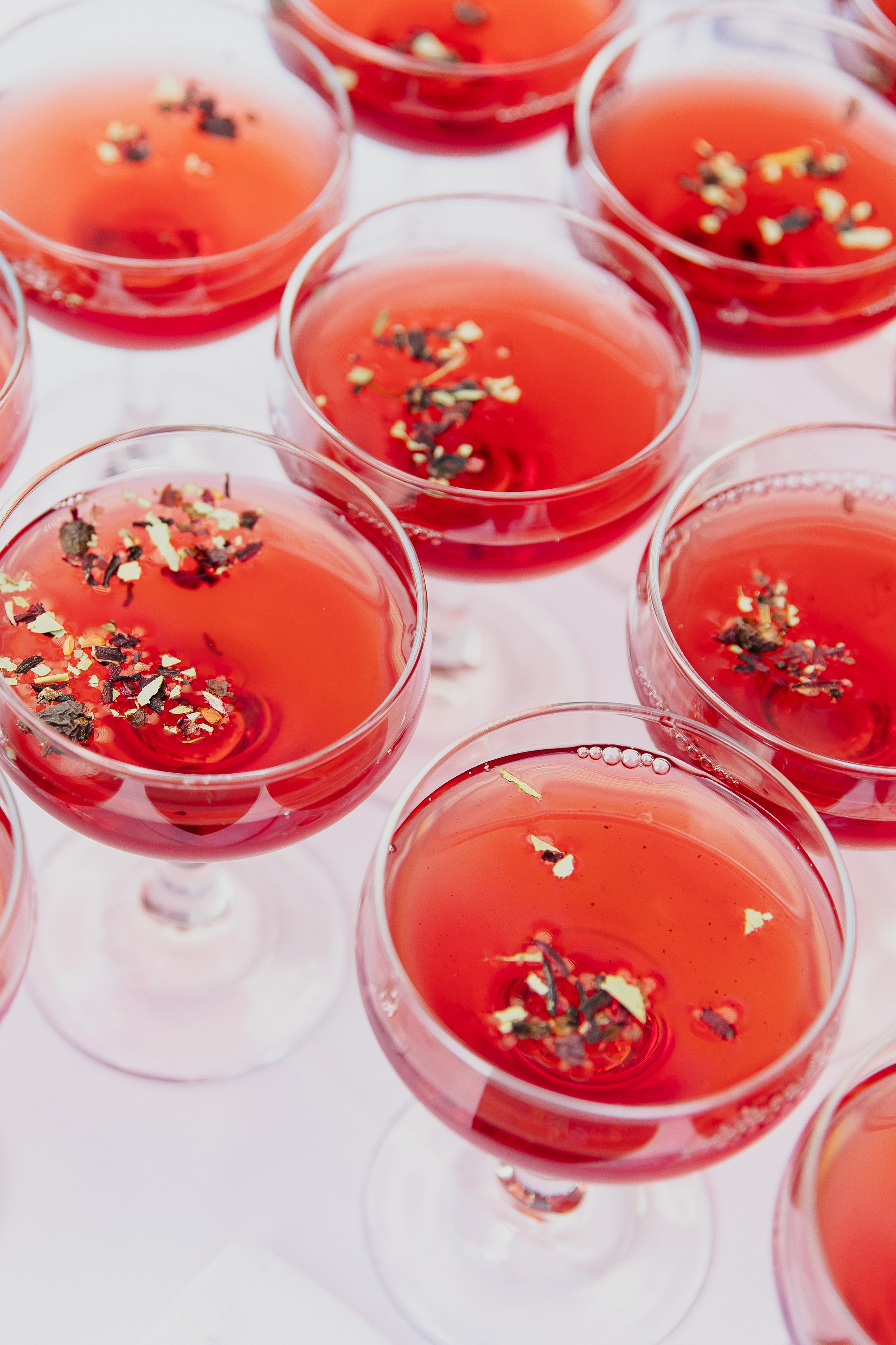 A high-angle view of multiple crystal coupe glasses arranged closely together, filled with a vibrant red cocktail. Each drink is garnished with a delicate sprinkle of dried loose tea leaves and herbs floating on the surface. The scene is softly lit against a white tablecloth background.