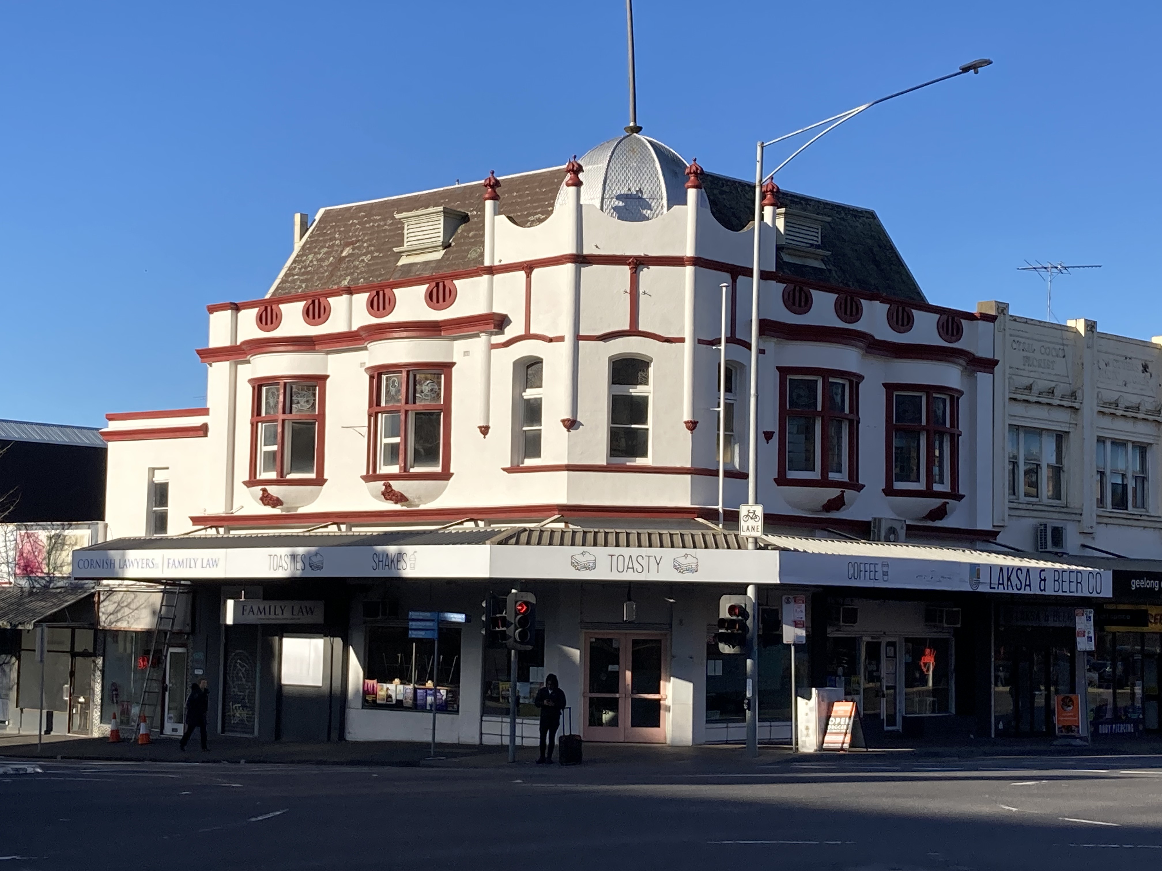 Heritage building on corner. White painted facade with maroon trim and sloping tiled roof.