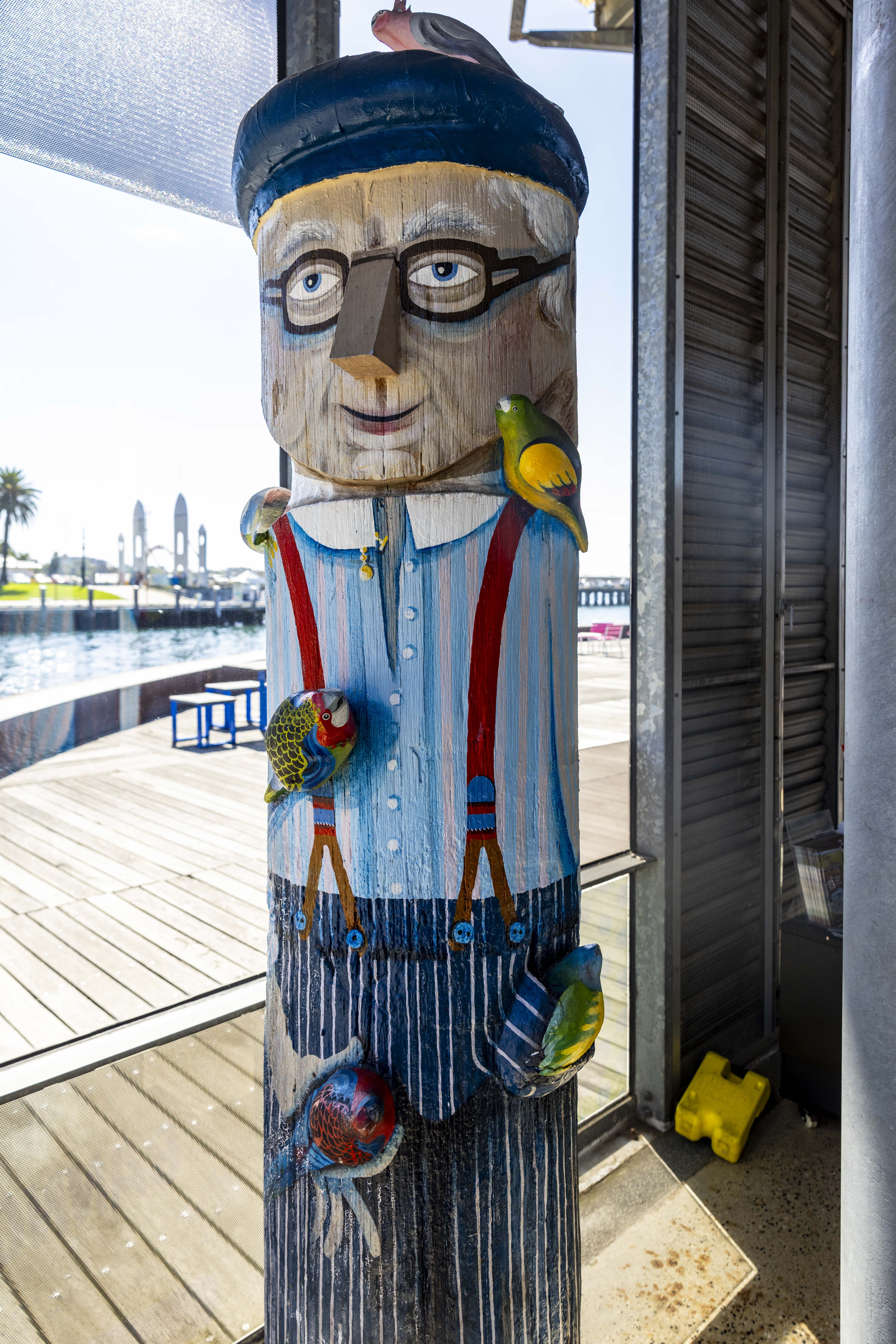 Painted bollard of local man with striped shirt, glasses, red suspenders and pants. His hat and clothes are covered in his colourful pet birds.