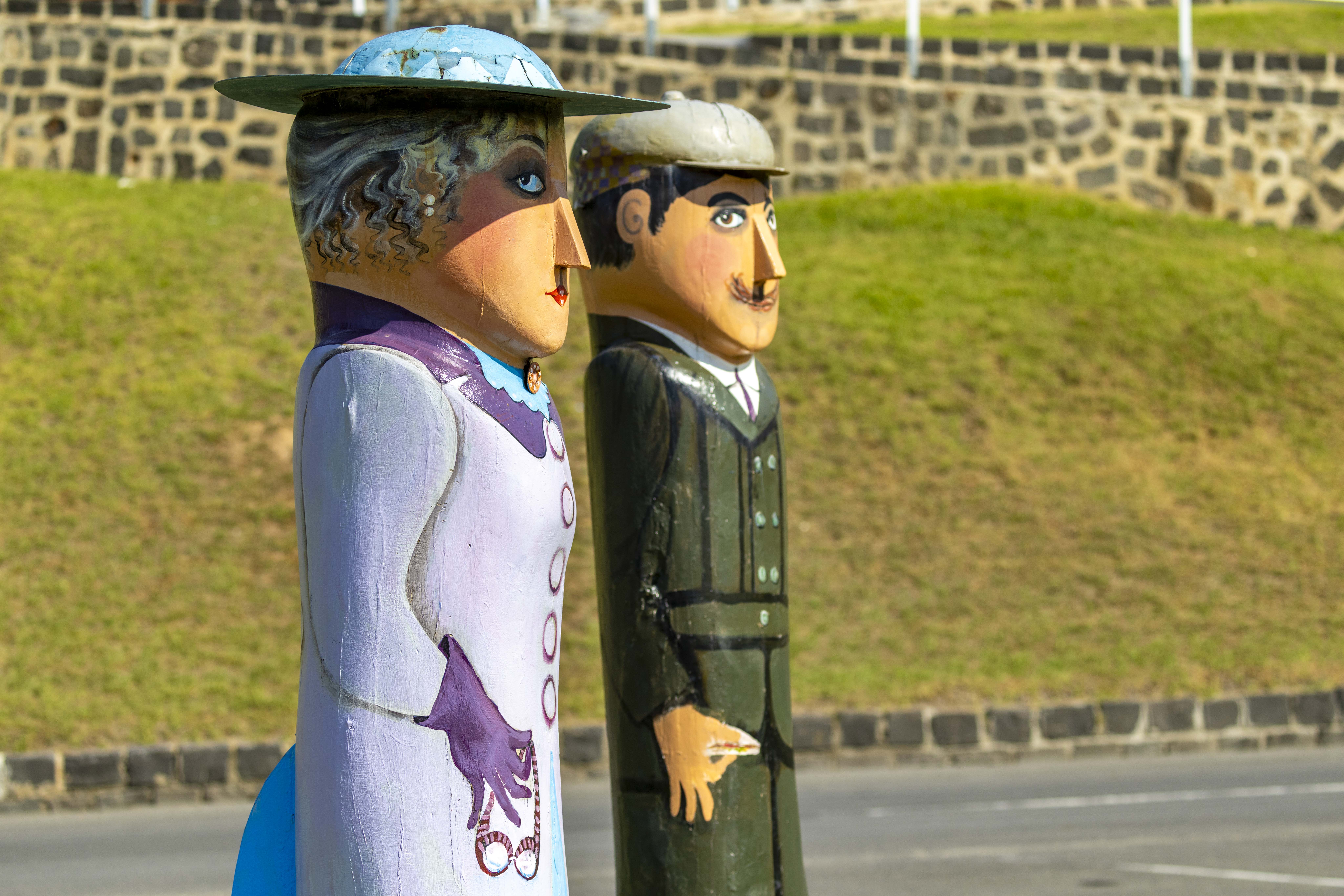 Two painted bollards of a husband and wife out for a weekend drive, dressed in formal weekend clothing. They are holding tools and driving googles which was the style at the time.