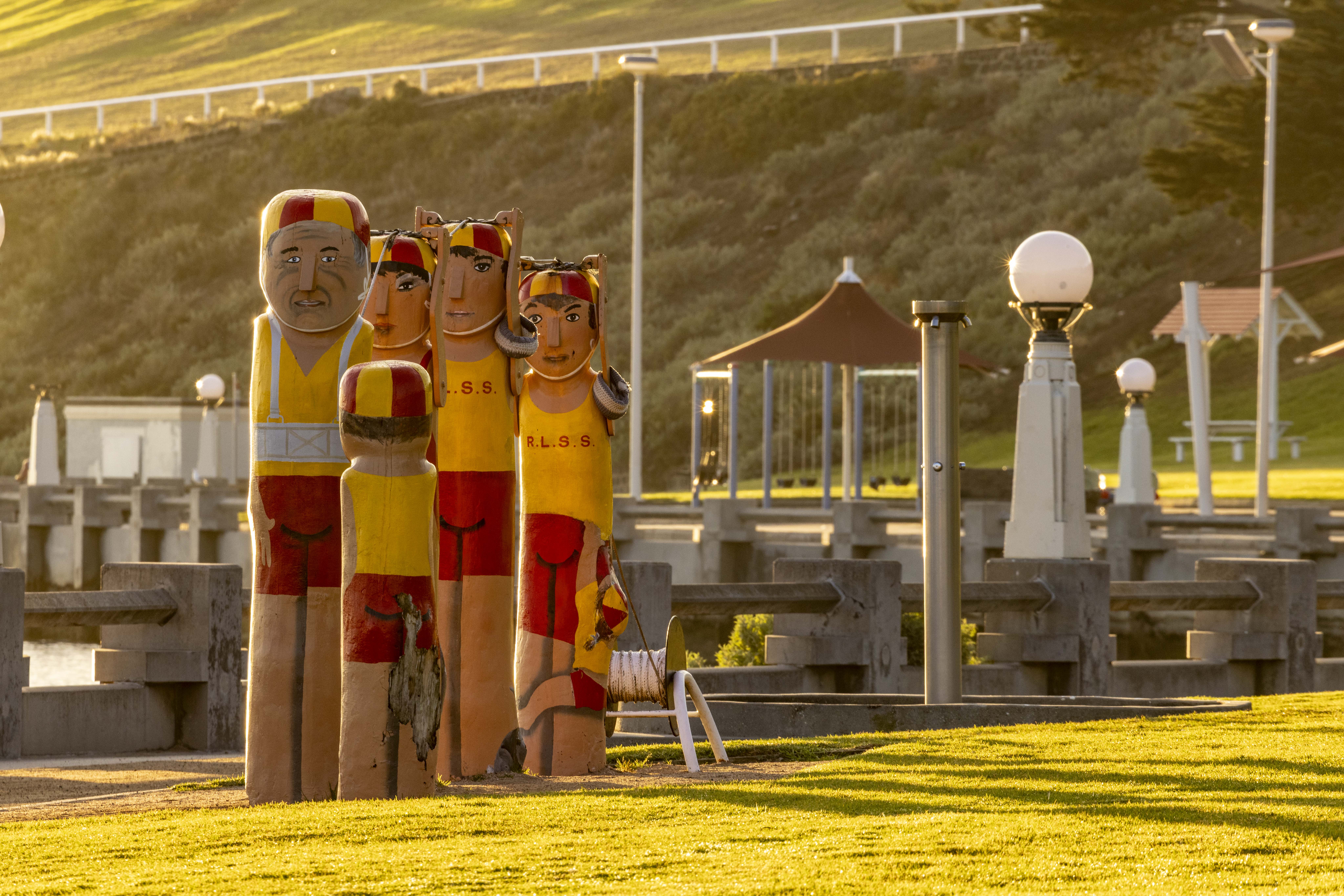 Five painted bollards dressed in traditional red and yellow lifesaving outfits. They are standing in a row and holding a rope above their heads.