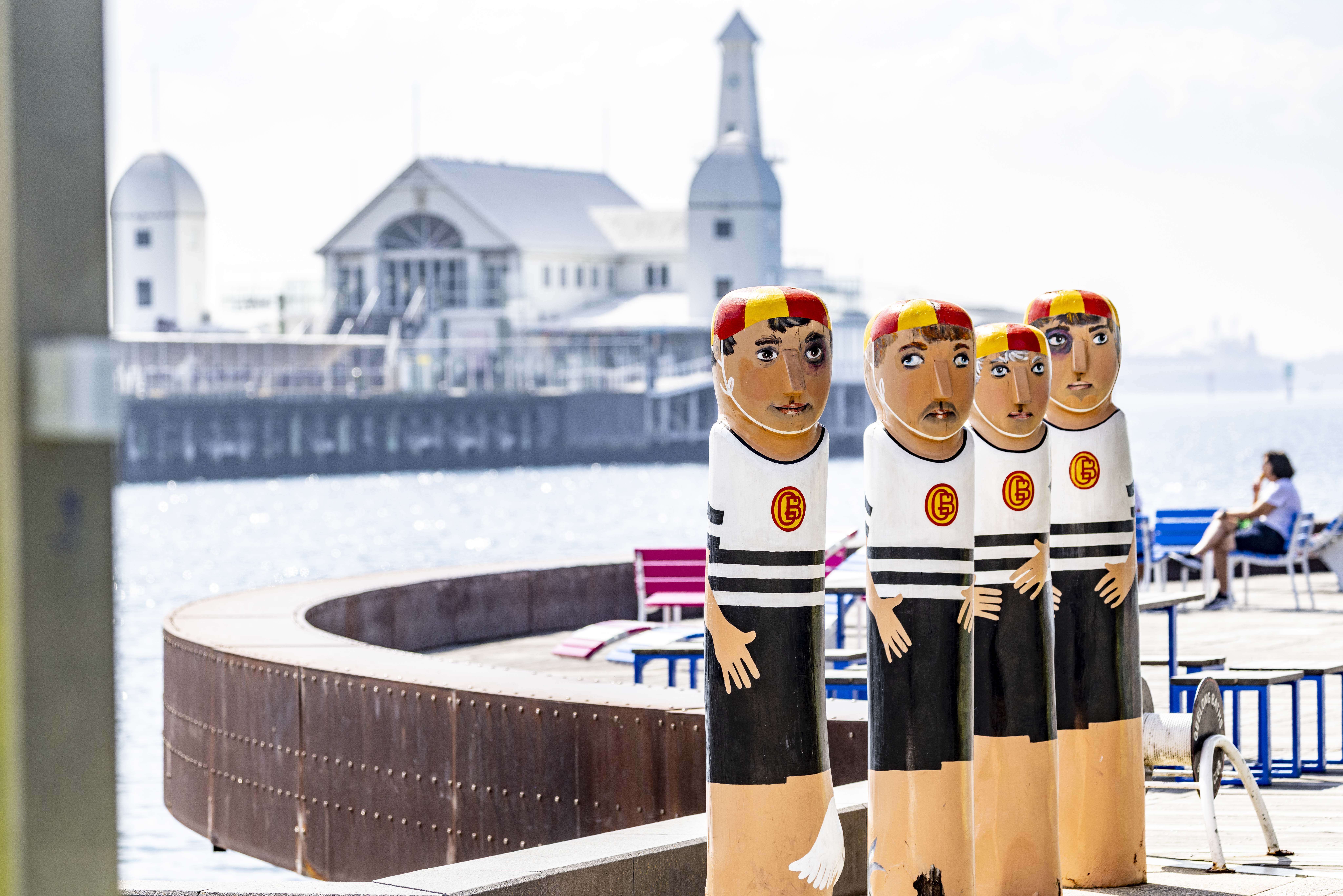 Four painted bollards of swimmers wearing white and blue swimsuits with yellow and red swim caps.