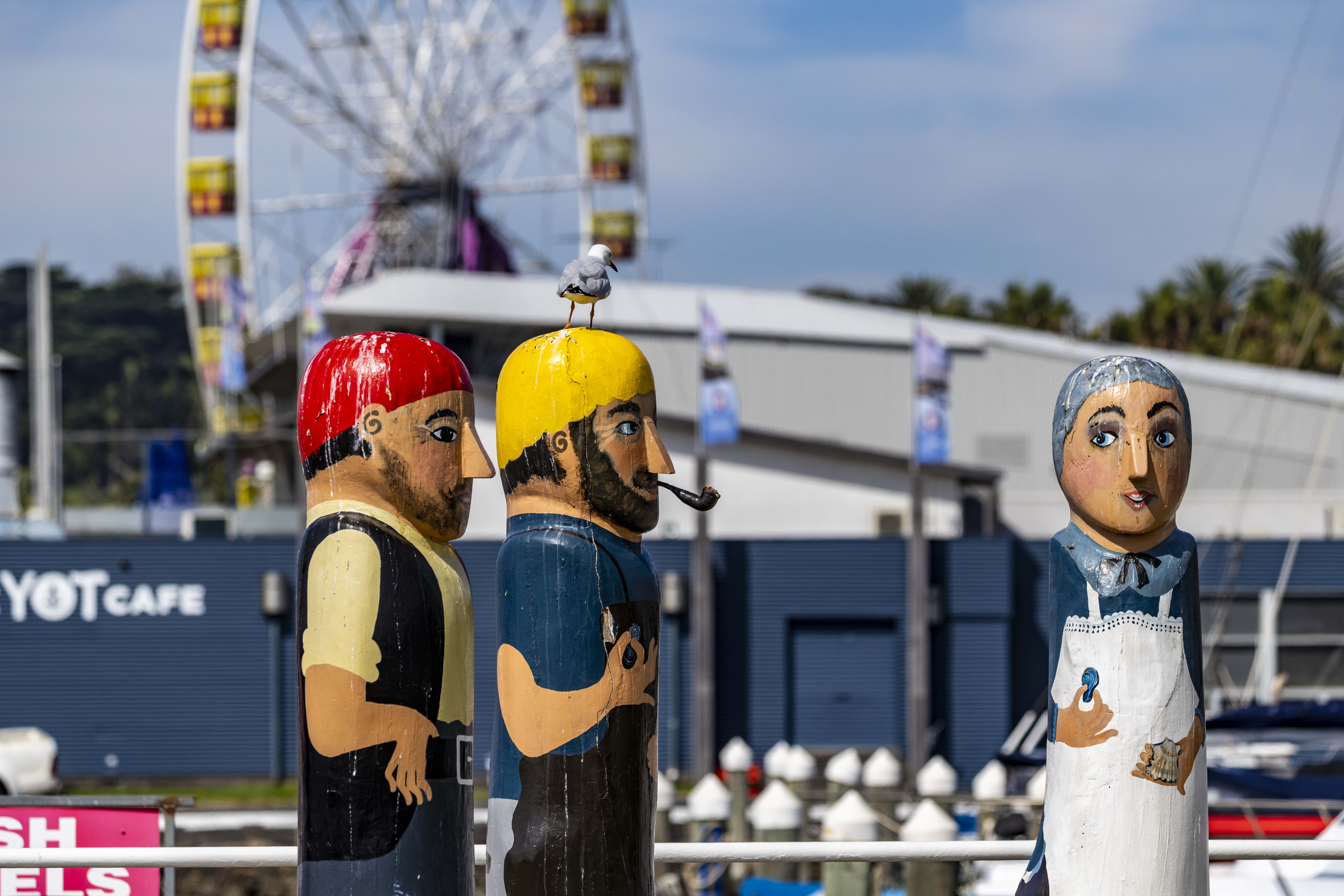 Three painted bollards of an old woman wearing a white apron and holding a scallop with two bearded fishermen. One has a red hat and the other has a yellow hat.