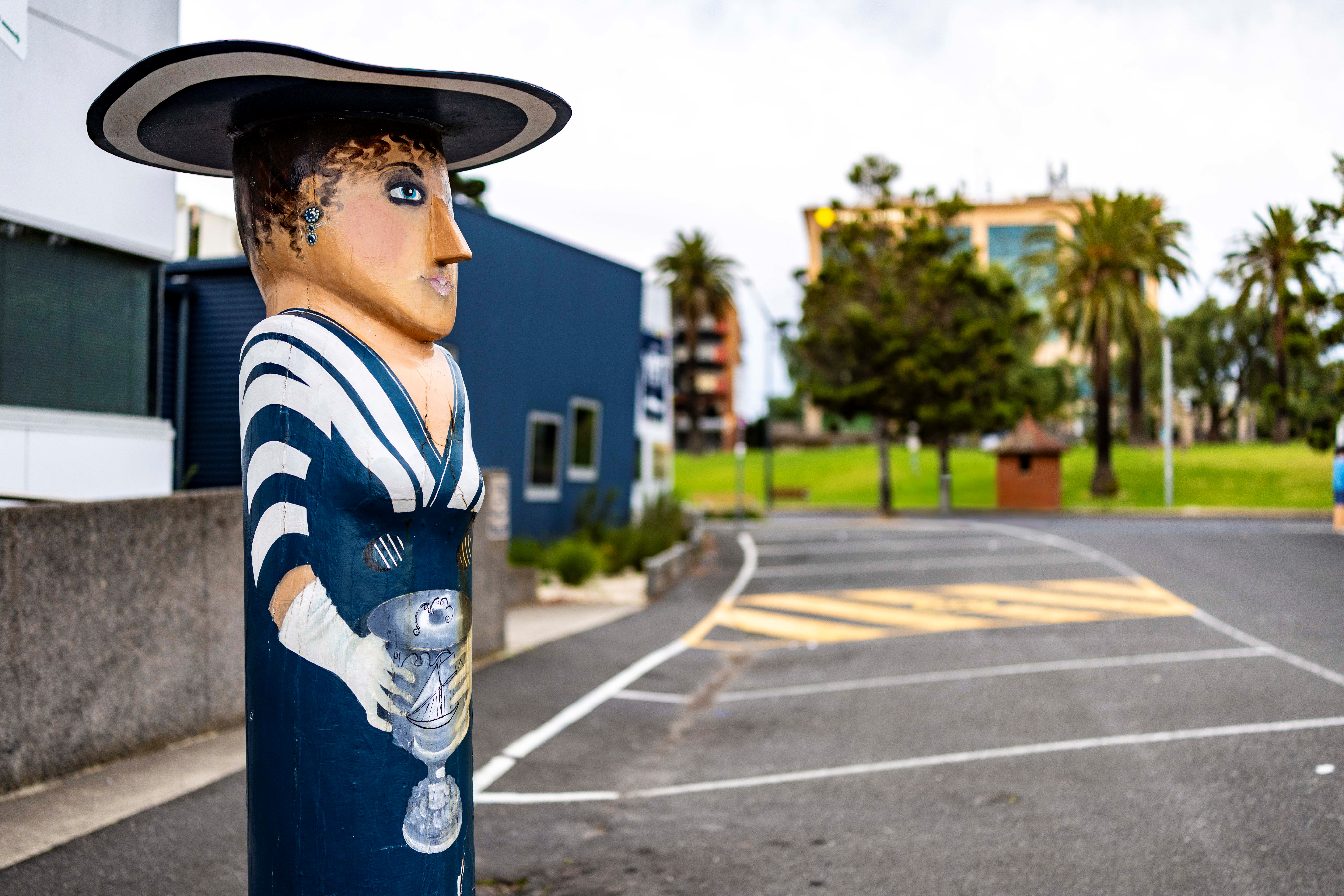 Painted bollard of a woman dressed in a blue and white striped dress and wide brimmed hat. She is standing out front of the Yacht Club and is holding a trophy.
