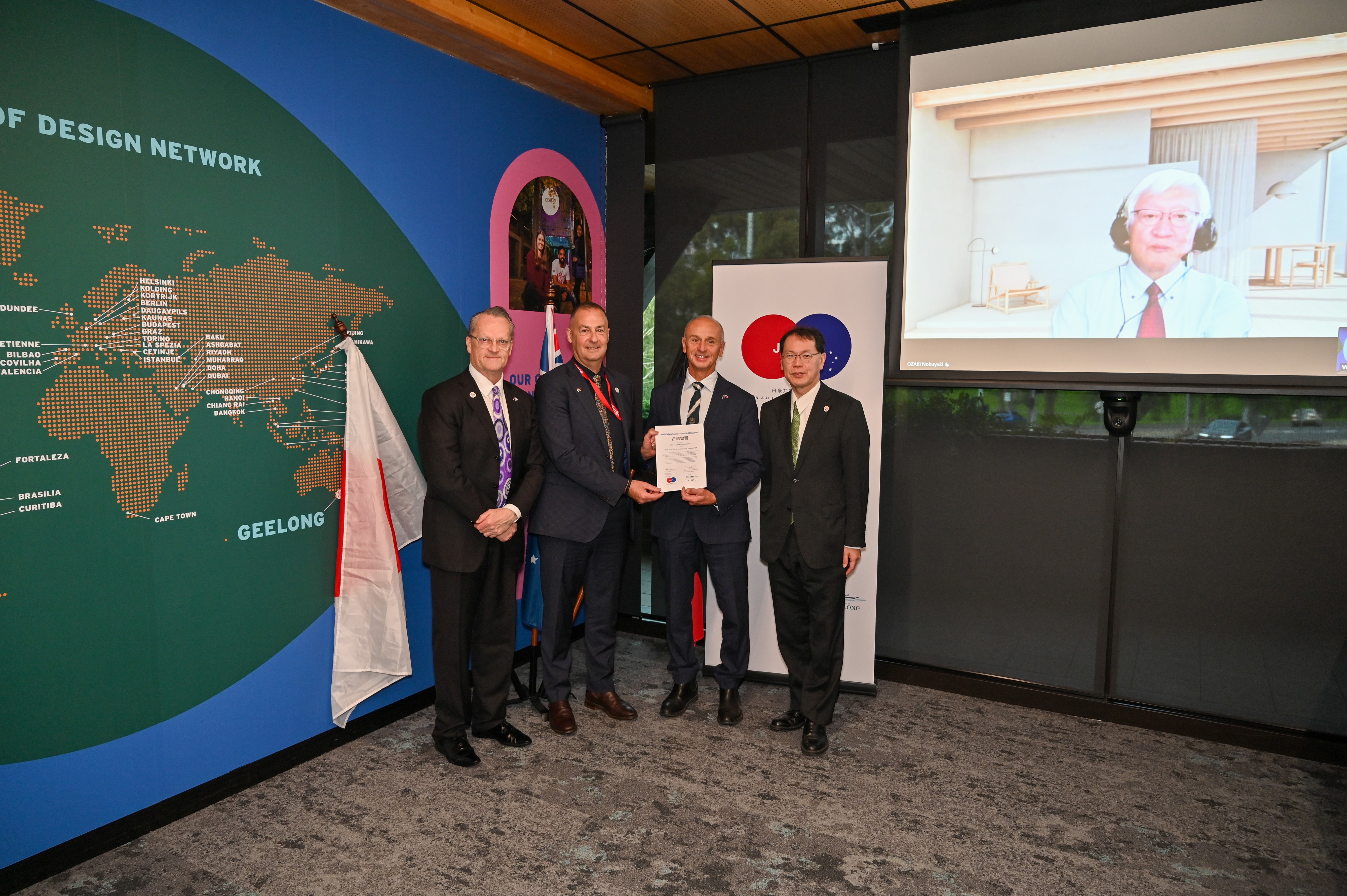 Four men standing holding a document with one person on screen in the background
