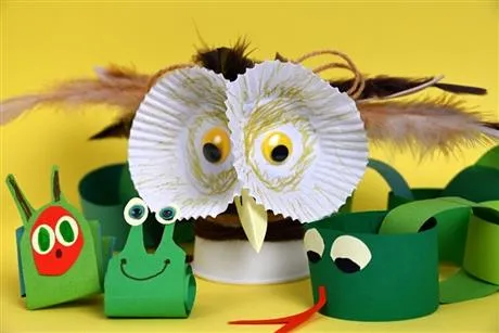 Colourful craft animals made from paper and cardboard, featuring an owl with large white eyes in the centre, surrounded by small green frog‑like figures on a yellow background.