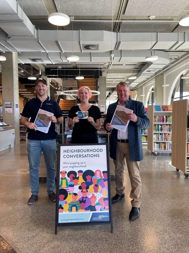 Three people standing indoors behind a freestanding sign that reads ‘Neighbourhood Conversations – We’re popping up in your neighbourhood’. Each person is holding an information flyer. Bookshelves and seating are visible in the background, indicating a public library or community space.