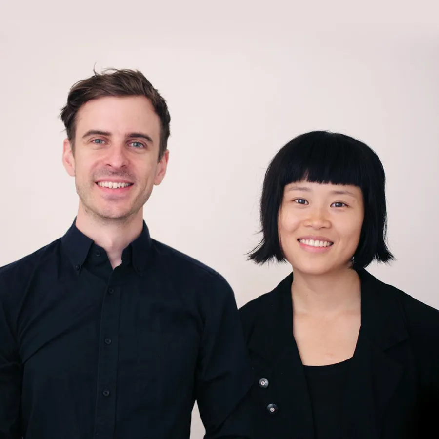 Two people standing side by side against a plain light background, wearing dark, professional-style clothing.
