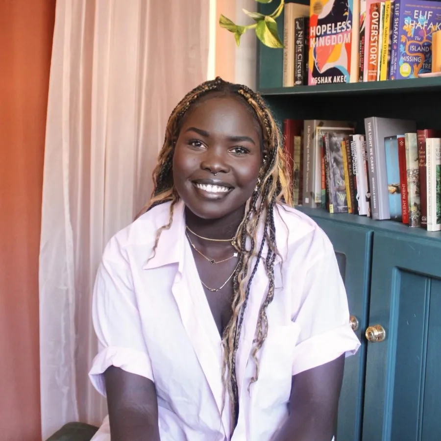 Person seated indoors in front of a bookshelf, wearing a white short‑sleeved shirt and layered necklaces, with long braided hair, in a softly lit room with books and a cabinet in the background.