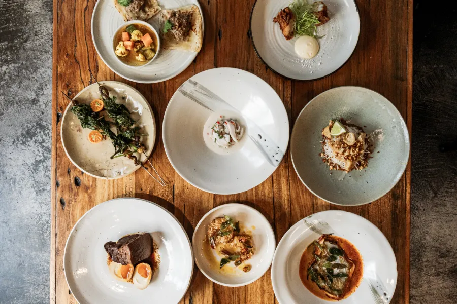 A flat lay overhead view of eight small plates filled with modern Asian fusion dishes arranged on a rustic wooden table. The selection includes skewered grilled greens with sliced kumquats, a pale soup bowl, braised meat with soft-boiled eggs, fried fish in a red curry sauce, and noodle dishes topped with crushed peanuts and lime.