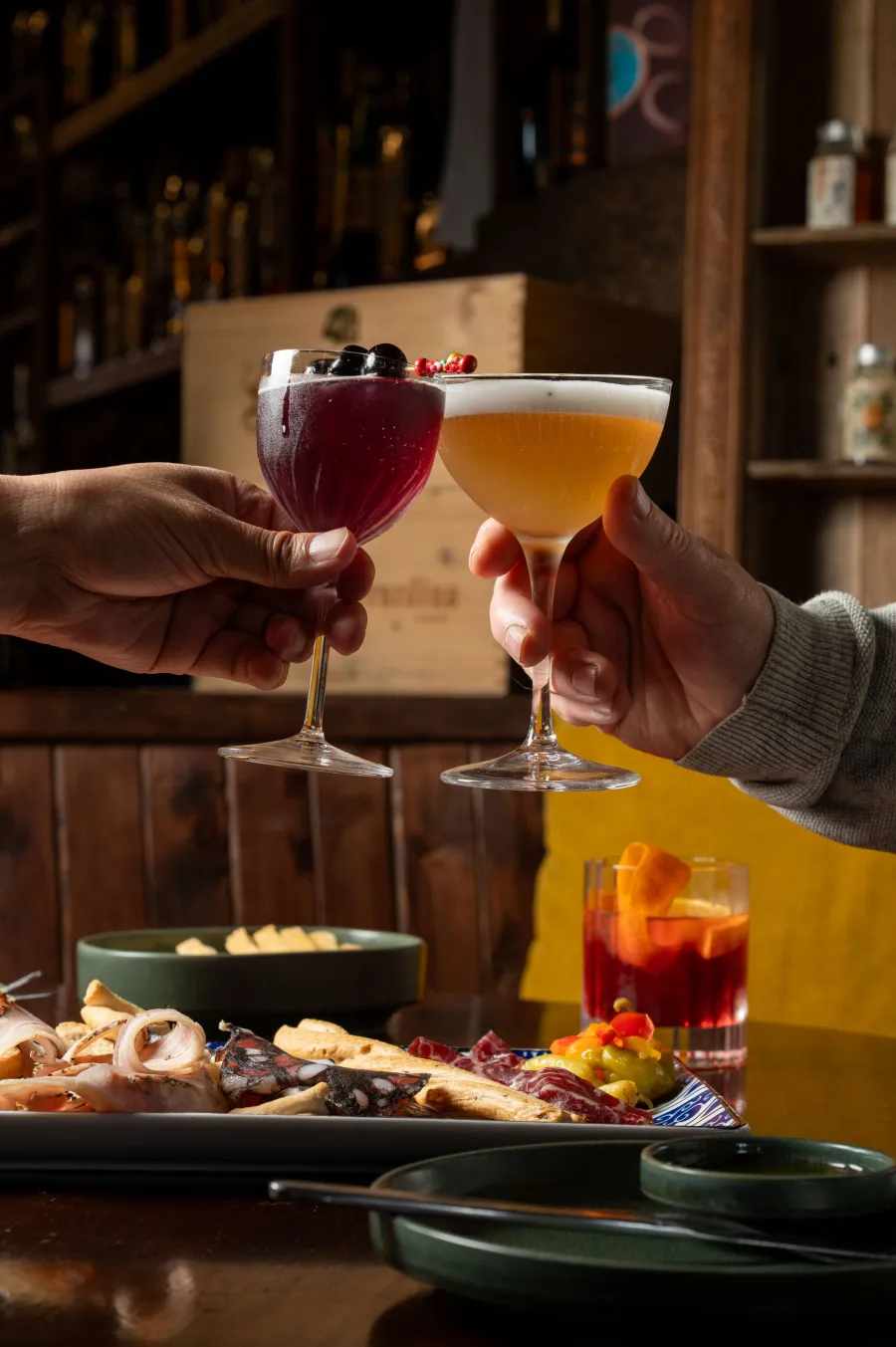 Two hands clinking cocktail glasses together over a charcuterie platter at a moody wood bar.