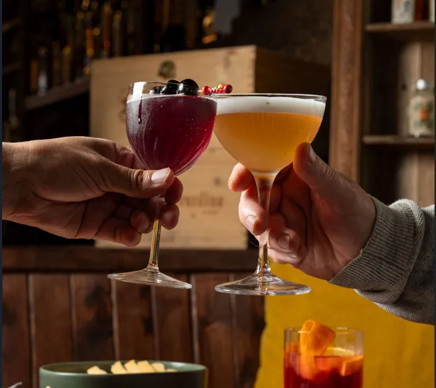 Two people clink elegant cocktails over a table filled with antipasto, cured meats, olives and flatbread in a warm, dimly lit bar setting. One drink is deep red with dark cherries as garnish, while the other is a pale frothy cocktail. A red aperitif sits in the background, creating a cosy and social atmosphere.