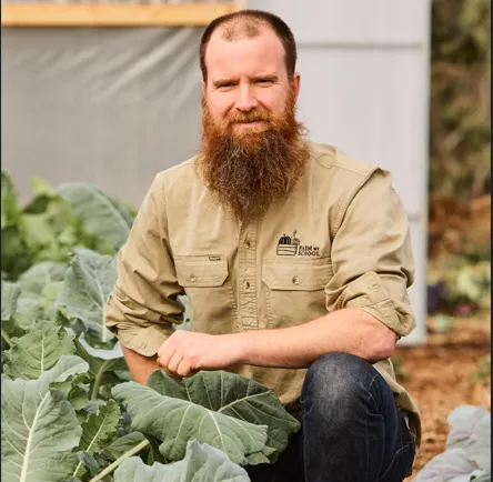 A man with a long red beard posing in a vegetable garden next to large green crops.