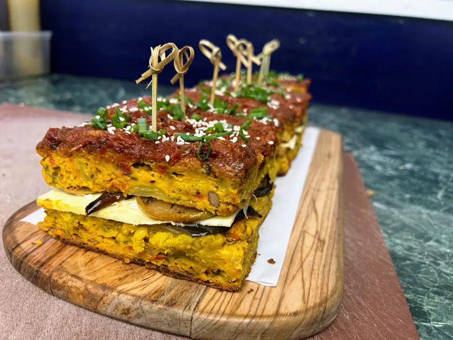 A row of savory zucchini frittata slices sandwiching a layer of cheese and roasted eggplant, served on a wooden board. The top layer is coated with a reddish tomato spread, sprinkled with sesame seeds and fresh chopped chives, and held together with decorative bamboo skewers.