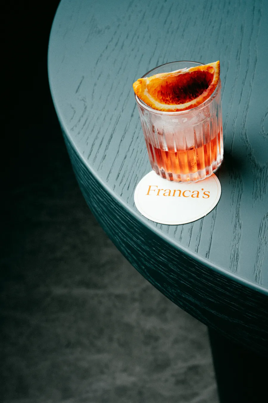 A Negroni cocktail with an orange slice sitting on a Franca’s coaster on a dark wooden table
