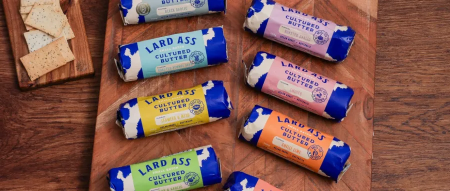 An overhead view of eight rolls of Lard Ass brand small-batch cultured butter arranged on a rustic wooden board. Each roll features bright blue and white packaging with distinct colorful labels indicating different flavors like smoked, roasted garlic, and chilli lime. A small wooden board with rectangular seed crackers sits in the upper left corner.