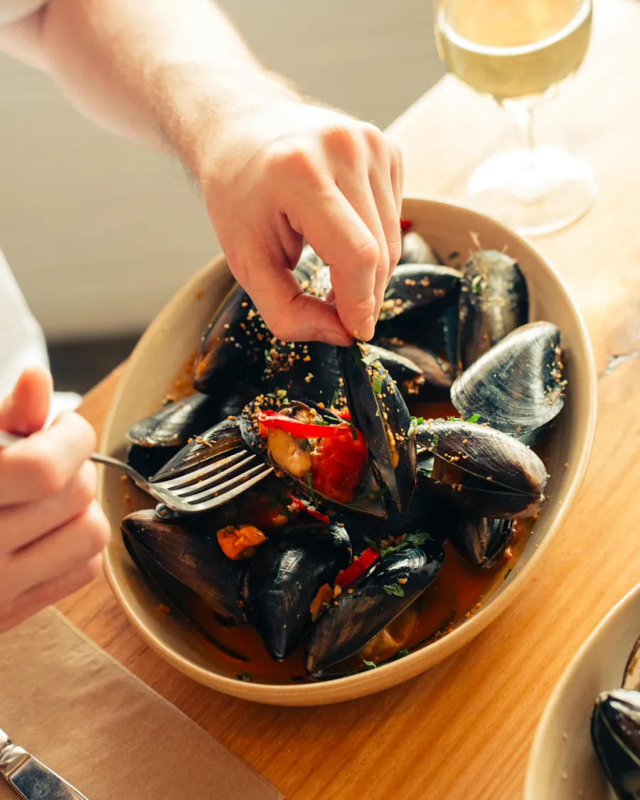 Top-down view of a person using their hands and a fork to eat steamed mussels from a large ceramic bowl. The mussels are served in a rich tomato broth with sliced red chili, fresh herbs, and a light seasoning crumble. A glass of white wine sits nearby on the wooden table.