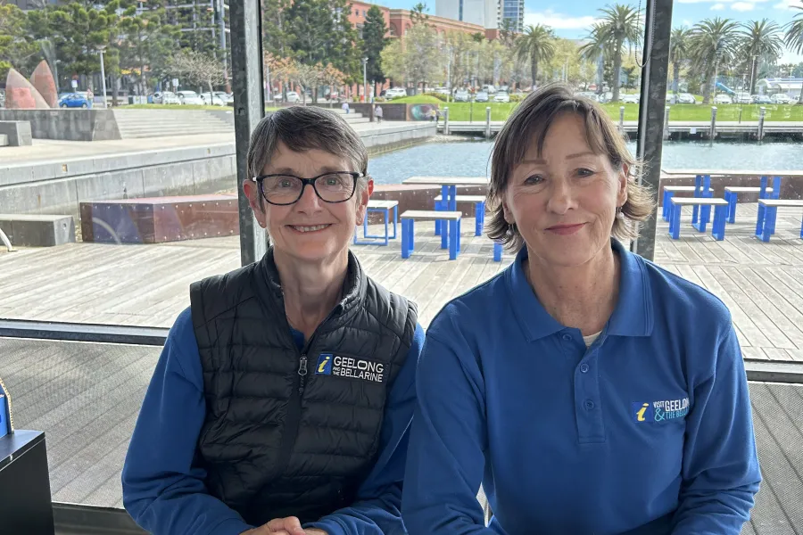 Volunteers Julie and Maureen Two Visit Geelong and The Bellarine volunteers seated at an information desk with brochures and maps, inside a glass-walled pavilion overlooking the Geelong waterfront and marina.
