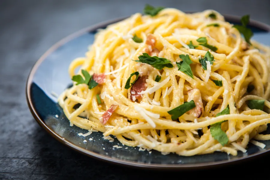 A plate of fresh spaghetti carbonara garnished with parsley and grated cheese on a dark background.