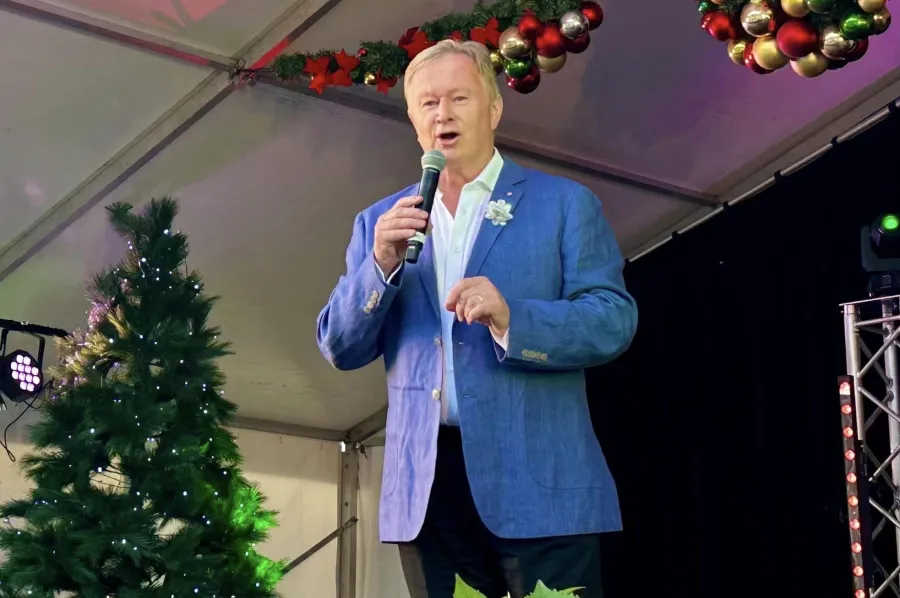 "Australian entertainer Denis Walter speaks into a microphone on an outdoor stage decorated with Christmas trees and festive baubles."