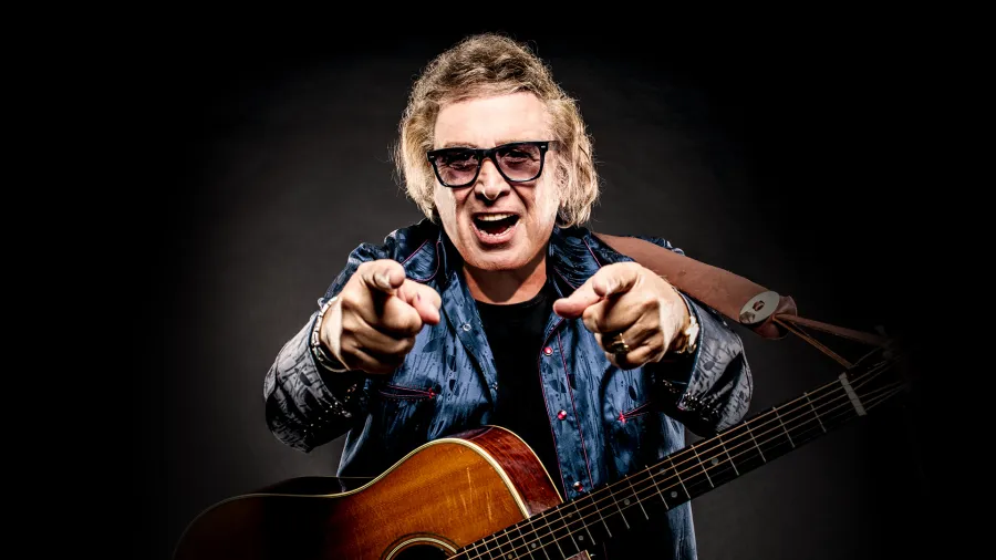 Don McLean posing with an acoustic guitar in front of black background.
