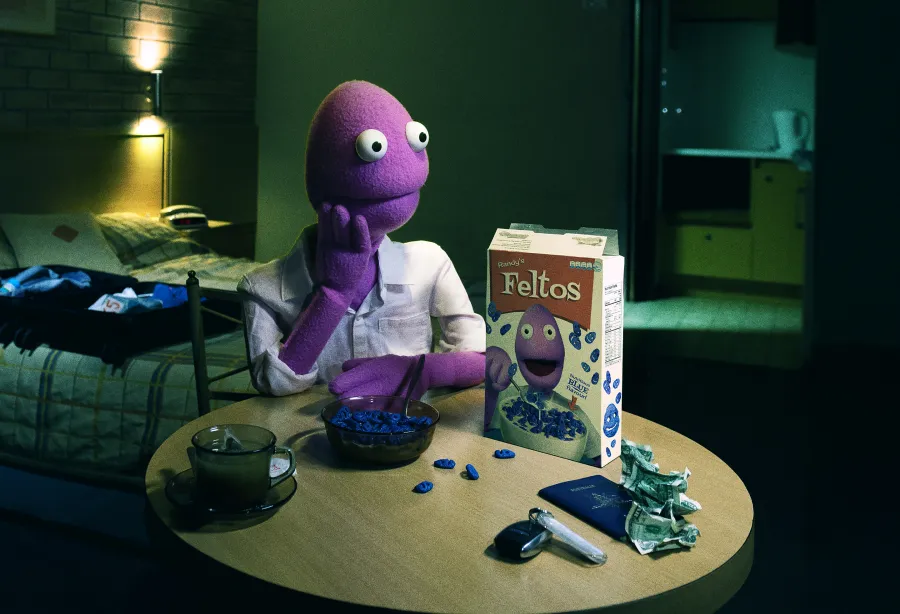 Randy Feltface puppet sitting at a dining table with a bowl of cereal labelled 'Feltos'