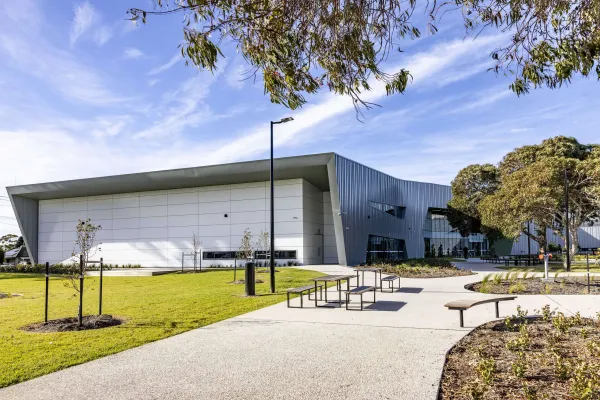 Exterior view of a modern community building with light grey and dark metal cladding, set beside a landscaped lawn. A paved pathway with benches and small trees leads to the entrance under a blue sky.