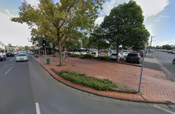 Brick-paved footpath with street trees, a bench, and a public phone on a corner beside a road with traffic lights showing green; parked cars fill a nearby car park, with low-rise shops lining the street in the background.