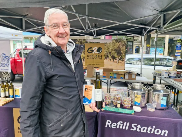 Steve Parker is a man with white hair and glasses and he stands in front of his market stall wearing a black raincoat and with one hand is holding a bottle and can of olive oil.