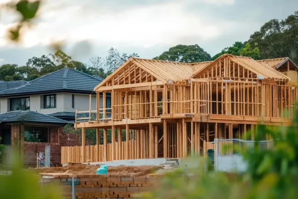 Two‑storey house under construction with exposed timber framing, next to completed homes and surrounded by trees.