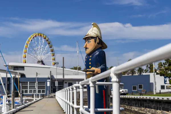 Fireman Painted bollard of a fireman, wearing a blue fireman uniform with gold painted fire helmet and holding a fire hose. The Geelong Ferris wheel and yacht club can be seen in the background.