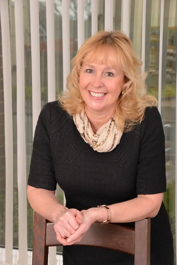 Artist Jan Mitchell Woman with black top and blonde hair leaning on the back of a wooden chair, smiling with hands clasped together.