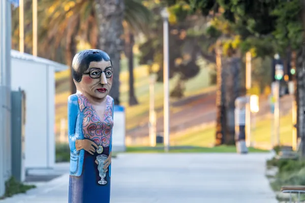 Mrs De Carteret Painted bollard of woman with glasses and hair tied back into a bun. She is wearing a floral shirt and blue cardigan and blue dress. She is holding a glass of lemonade and a slice of lemon.