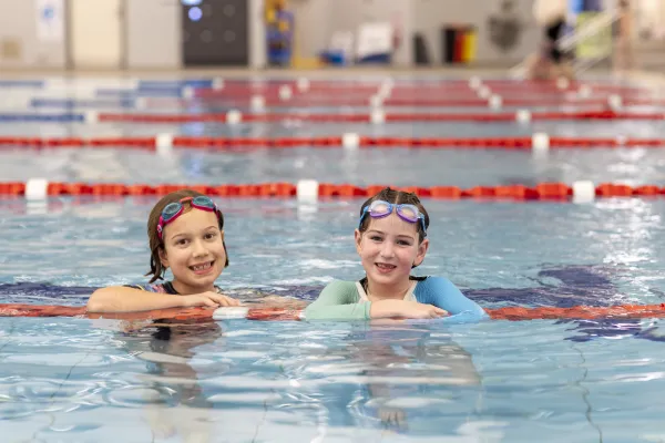 Two young swimmers with goggles on their heads are in the pool leaning on a lane rope and smiling.