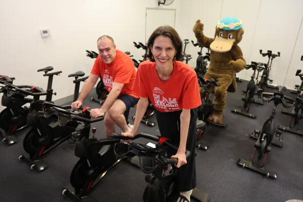 Craig Williams, Ali Wastie and Webstar Swim School mascot riding exercise bikes