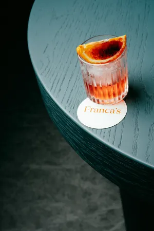 A Negroni cocktail with an orange slice sitting on a Franca’s coaster on a dark wooden table