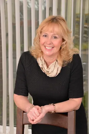 Woman with black top and blonde hair leaning on the back of a wooden chair, smiling with hands clasped together.